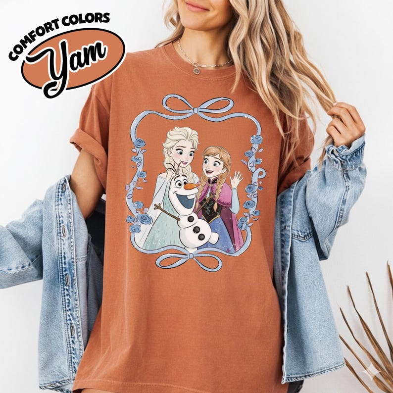May include: A rust-colored t-shirt featuring a cartoon image of Elsa, Anna, and Olaf from the movie Frozen, framed by a blue floral design. The shirt has the words "COMFORT COLORS" and "Yam" in a brown oval.