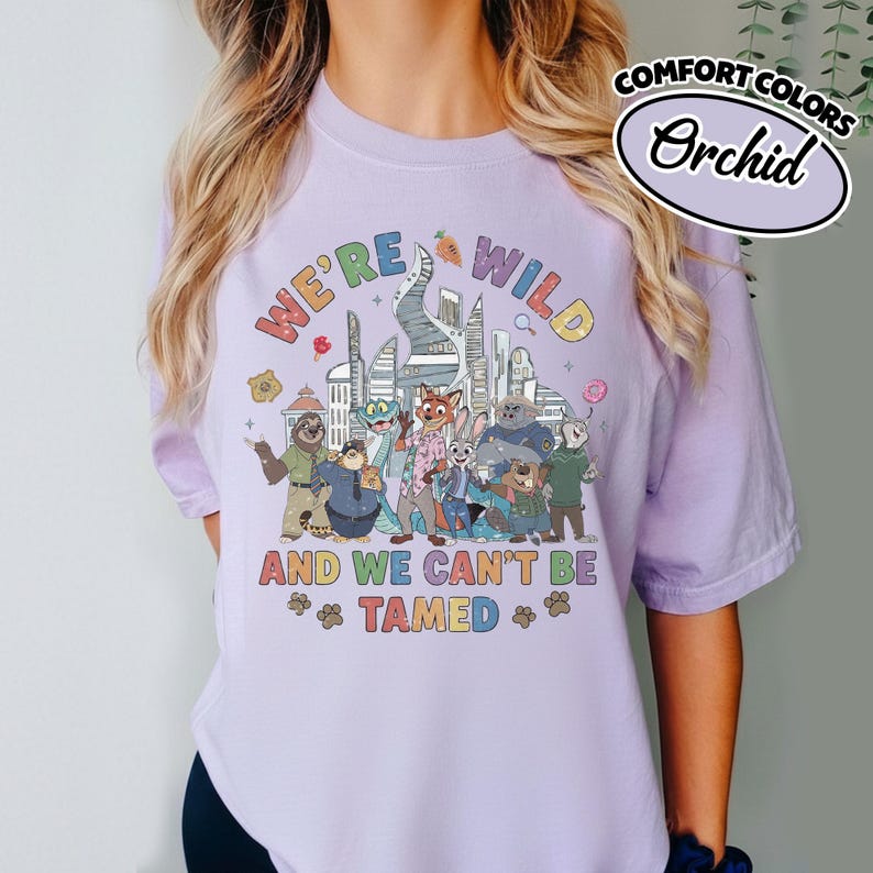 May include: A light purple t-shirt with the text "WE'RE WILD AND WE CAN'T BE TAMED" and a colorful graphic of animal characters in front of a city skyline. The shirt is labeled "Comfort Colors" and the color is "Orchid".