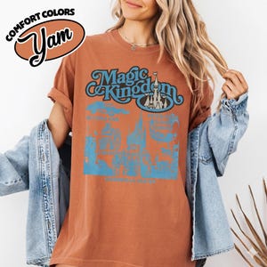 May include: A rust-colored t-shirt with the words "Magic Kingdom" in blue script and a castle graphic. The shirt also features additional blue graphics and text, with the Comfort Colors and Yam logos in the upper left corner.