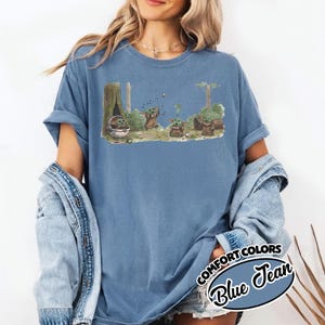 May include: Blue t-shirt with a graphic of a character from a popular science fiction series. The design features the character in a forest setting. The shirt is labeled "Comfort Colors Blue Jean".