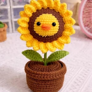 May include: A handcrafted crochet sunflower with a smiling chick face in a brown pot. The sunflower has yellow petals, a brown center, and green leaves. The pot is also brown. A charming, whimsical home decor item.
