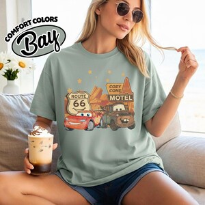 May include: Sage green t-shirt featuring a graphic of characters from the movie Cars, including Lightning McQueen and Mater, with Route 66 and Cozy Cone Motel signs. The shirt has the Comfort Colors Bay logo.