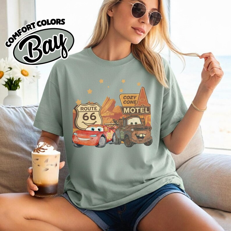 May include: Sage green t-shirt featuring a graphic of characters from the movie Cars, including Lightning McQueen and Mater, with Route 66 and Cozy Cone Motel signs. The shirt has the Comfort Colors Bay logo.