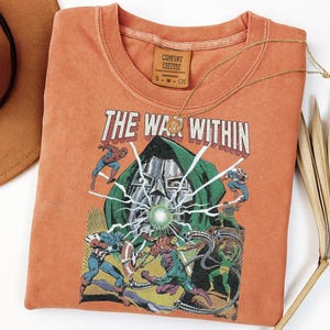 May include: Orange t-shirt featuring a comic book graphic with the text "THE WAR WITHIN." The graphic depicts various superheroes in battle. A gold necklace with a pendant rests on the shirt. The shirt has a Comfort Colors tag.