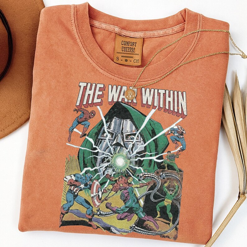 May include: Orange t-shirt featuring a comic book graphic with the text "THE WAR WITHIN." The graphic depicts various superheroes in battle. A gold necklace with a pendant rests on the shirt. The shirt has a Comfort Colors tag.