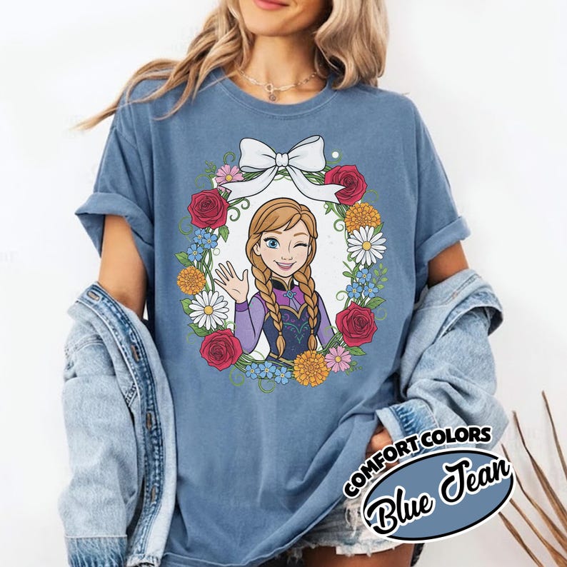 Peut inclure: T-shirt bleu avec une image de dessin anim&eacute; d'un personnage aux cheveux tress&eacute;s, faisant un clin d'&oelig;il et saluant, entour&eacute; d'une couronne florale et d'un n&oelig;ud blanc. Le t-shirt est &eacute;tiquet&eacute; "Comfort Colors Blue Jean".