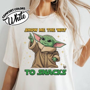 May include: White Comfort Colors t-shirt featuring a green cartoon character with large ears, pointing upwards. The text "SHOW ME THE WAY TO SNACKS" is printed in yellow and green. The shirt has a round neckline.