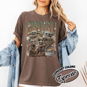K&ouml;nnte beinhalten: Ein braunes Kurzarm-T-Shirt mit einem Grafikdruck der Figur Grogu in verschiedenen Posen, einschlie&szlig;lich einer in einer schwebenden Kapsel. Der Text GROGU ist in gro&szlig;en gr&uuml;nen und wei&szlig;en Buchstaben &uuml;ber den Illustrationen abgedruckt.
