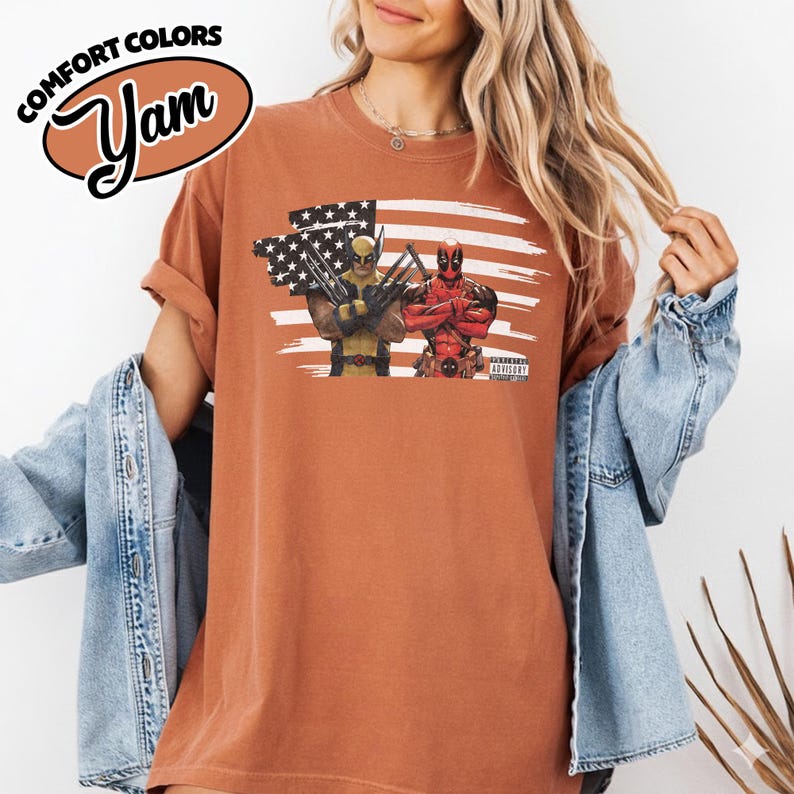 May include: A rust-colored short-sleeve t-shirt featuring a graphic print of Wolverine and Deadpool standing in front of a stylized American flag. The graphic includes the text Parental Advisory. The shirt has a crew neckline and a relaxed fit.