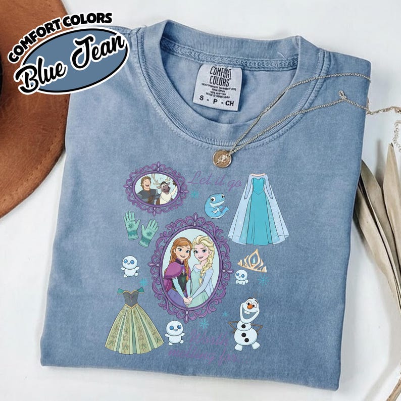 Op de afbeelding: Blauw spijkerbroek Comfort Colors T-shirt met een ontwerp uit de film "Frozen". Het ontwerp bevat afbeeldingen van Anna en Elsa, jurken en de zin "Let it go".