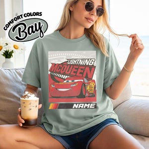 May include: A sage green t-shirt featuring a graphic print of the red race car Lightning McQueen with the text Lightning McQueen and Name. The design includes a speedometer background and a red, orange, and yellow stripe accent at the bottom.