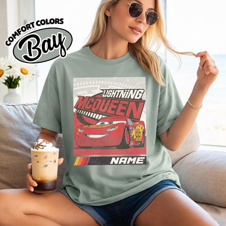 May include: A sage green t-shirt featuring a graphic print of the red race car Lightning McQueen with the text Lightning McQueen and Name. The design includes a speedometer background and a red, orange, and yellow stripe accent at the bottom.