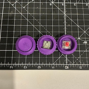 May include: Three purple, 3D-printed mechanical keyboard components are arranged on a black cutting mat. The components include a solid cap, a switch housing with a white switch, and a switch housing with a red switch.