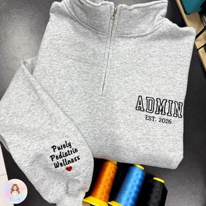 Puede incluir: Una sudadera gris con cremallera de cuarto de cuello con la palabra "ADMIN" y "EST. 2026" bordada en la parte delantera. La manga izquierda tiene bordado "Purely Pediatric Wellness" con un corazón rojo. Se ven carretes de hilo.