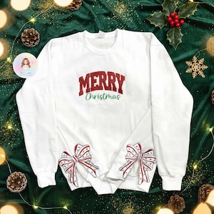 Pode incluir: Camisola branca com a inscrição "MERRY Christmas" em vermelho e verde. A camisola apresenta dois laços bordados com riscas vermelhas e brancas. Roupa festiva para a época natalícia.