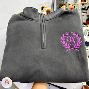 Pode incluir: Um moletom cinza carvão com gola com zíper e um emblema magenta bordado no peito esquerdo. O emblema apresenta um monograma dentro de um design de coroa de louros. O moletom é feito de um material macio e espesso.