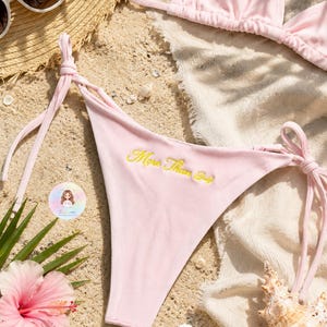 May include: A light pink ribbed bikini bottom featuring side tie closures and the yellow embroidered text More Than 24. The garment is crafted from a textured fabric and includes thin adjustable straps on both sides for a secure fit.
