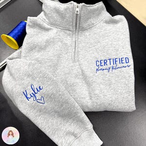 May include: A gray quarter-zip sweatshirt with blue embroidered text that reads "CERTIFIED pharmacy technicians" and "Kylie" with a heart. The sweatshirt has a high collar and a silver zipper.