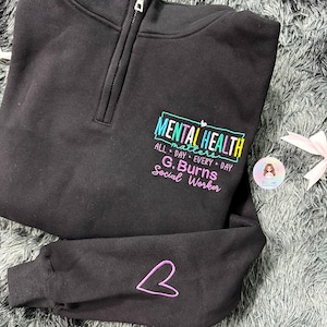 Könnte beinhalten: Ein schwarzes Sweatshirt mit kurzem Reißverschluss und einer bunten Stickerei mit dem Text MENTAL HEALTH matters ALL DAY EVERY DAY G. BURNS Social Worker. Der Ärmel ist im Manschettenbereich mit einer violetten Herzumrandung bestickt.