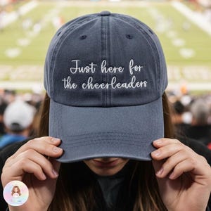 以下が含まれることがあります： 「Just here for the cheerleaders」の文字が白い刺繍で施された、ネイビーブルーの野球帽。カーブしたつばと、カジュアルな装いに適したクラシックな6パネルのデザインです。