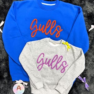 Könnte beinhalten: Ein leuchtend blaues Sweatshirt mit Rundhalsausschnitt und orangefarbenem kursivem Schriftzug Gulls sowie ein kleineres graues Sweatshirt mit violettem kursivem Schriftzug Gulls und einer kleinen gelben Schleife am Halsausschnitt.