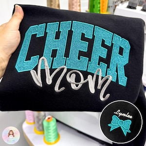 May include: A black sweatshirt featuring the word CHEER in large, teal glittery block letters with the word Mom embroidered below in a silver cursive script. A circular inset shows a teal glittery bow and the name Lynlee in white script.