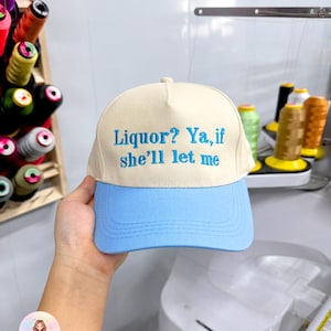 May include: A beige and light blue baseball cap with the embroidered text "Liquor? Ya, if she'll let me". The cap has a curved brim and a structured crown. The text is in a light blue color.