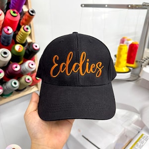 Può includere: Cappellino da baseball nero con il nome "Eddies" ricamato in filo arancione. Il cappellino ha una visiera curva ed è realizzato in tessuto resistente. Lo sfondo mostra rocchetti di filo colorato.