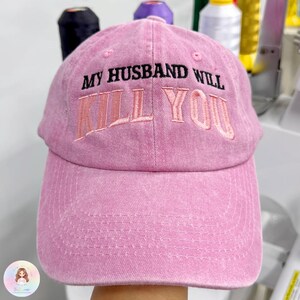 May include: A pink baseball cap with the embroidered text "MY HUSBAND WILL KILL YOU" in black and pink thread. The cap has a curved brim and a faded, vintage look.