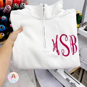 May include: A white quarter-zip sweatshirt featuring a silver-toned metal zipper at the collar and a large, dark pink embroidered monogram with the letters MSB in a decorative cursive script on the lower right side of the chest area.