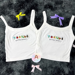 May include: Two white vest tops with thin straps. Each top features an embroidered patch with various bugs and the text "sad bug summer." Yellow, purple, and pink bows are scattered around.