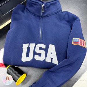 Peut inclure: Sweat-shirt bleu marine avec un col montant et une fermeture éclair quart de zip. Le mot "USA" est brodé en blanc sur la poitrine. Un écusson du drapeau américain est sur la manche gauche.