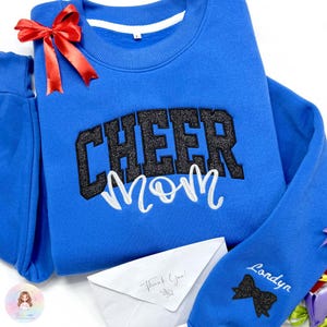 Peut inclure: Un sweat-shirt bleu avec les mots "CHEER MOM" en paillettes noires et en écriture blanche. Un nœud rouge est attaché au sweat-shirt. Un pantalon de survêtement bleu assorti et une enveloppe blanche avec "Thank You!" sont également visibles.