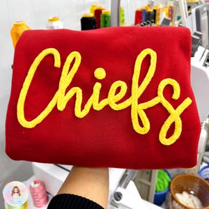 May include: A red sweatshirt featuring the word "Chiefs" in yellow embroidered script. The garment is made of a soft material, likely a fan item.