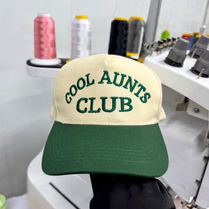 Op de afbeelding: Crème en groene baseballcap met de geborduurde tekst "COOL AUNTS CLUB". De cap wordt getoond tegen een achtergrond met naaimachines en spoelen garen. Een modieus accessoire.