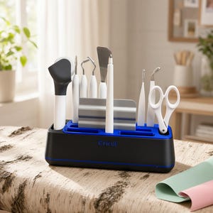 Puede incluir: Un organizador de herramientas Cricut negro y azul que contiene varios utensilios de manualidades, incluyendo un cortador rotativo, un raspador de metal, un gancho de depilar, pinzas y tijeras de mango blanco en una base rectangular azul.