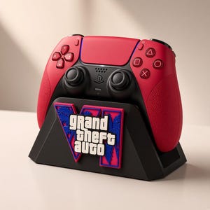 以下が含まれることがあります： 黒い充電スタンドに置かれた赤と黒のワイヤレスビデオゲームコントローラー。スタンドには、青とピンクのヤシの木のシルエットが描かれたGrand Theft Autoのロゴが目立つように配置されています。コントローラーには標準的なボタンとジョイスティックが付いています。