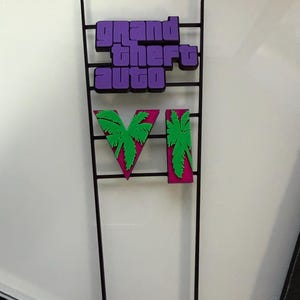 Könnte beinhalten: Eine schwarze Metallrahmenstruktur mit dem violetten Schriftzug "grand theft auto", der über der römischen Ziffer "VI" in Pink mit grünen Palmensilhouetten auf jedem Zeichen angeordnet ist, montiert auf zwei vertikalen schwarzen Metallstangen.