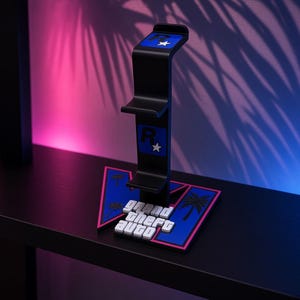 Op de afbeelding: Een zwarte verticale gaming headset standaard met het Rockstar Games logo in blauw en wit aan de bovenkant en in het midden. De basis is een blauwe en roze driehoekige vorm met palmbomsilhouetten en de witte tekst Grand Theft Auto.