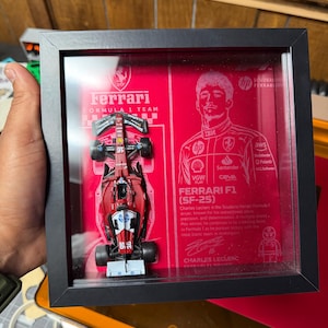 Puede incluir: Un marco de caja negra que contiene un modelo de coche de carreras Ferrari de Fórmula 1 rojo y una tarjeta de presentación roja con un retrato de Charles Leclerc, el texto Ferrari F1 SF-25, logotipos, una firma impresa y un pequeño personaje.
