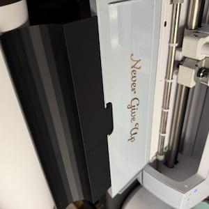 Puede incluir: Una máquina de corte Cricut Maker 3 blanca con un panel lateral negro. La máquina cuenta con una tapa blanca con el texto 'Never Give Up' impreso en una fuente cursiva dorada metálica. El carro interno sostiene dos soportes A y B.