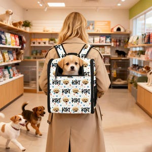 May include: A tan and white dog peeks out of a dog carrier backpack. The carrier has a white background with a pattern of dog faces, paw prints, and the name "KIKI" in black. The person is wearing a tan trench coat.