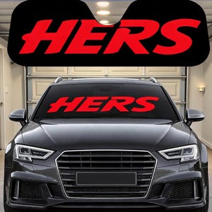 May include: A black car windshield sun shade featuring the word HERS printed in large, bold red capital letters. The sun shade is positioned against the front windshield of a dark gray vehicle, displaying the text clearly across the center.