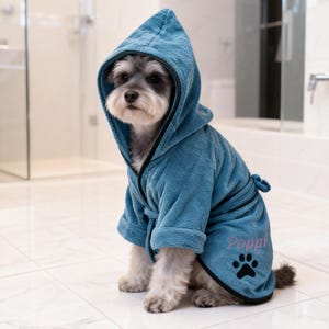 Peut inclure: Un petit chien portant un peignoir à capuche bleu sarcelle avec une bordure noire. Le peignoir a une ceinture, le nom "Poppi" en script rose et une empreinte de patte noire. Le chien est assis sur un sol carrelé blanc.