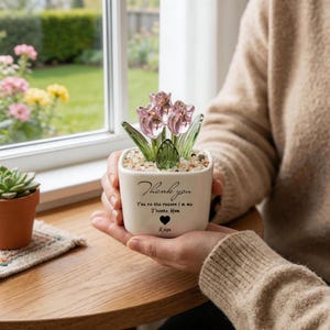 May include: A small, square, off-white ceramic pot with a glass flower arrangement. The arrangement features pink tulips and green leaves. The pot has the words "Thank you" and "You're the reason I'm me. Thanks Mom." written on it.