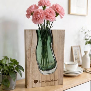May include: A wooden vase with a green glass insert holding pink carnations. The vase has the text "Love you, Mom!" and a heart symbol engraved on the wood. The vase is on a wooden surface.