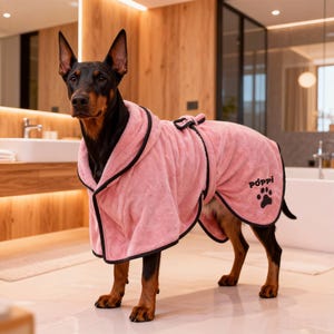 May include: A Doberman Pinscher wearing a pink terry cloth dog robe with black trim. The robe has a hood and a bow detail on the back. The word "Pöppi" and a paw print are embroidered on the robe.