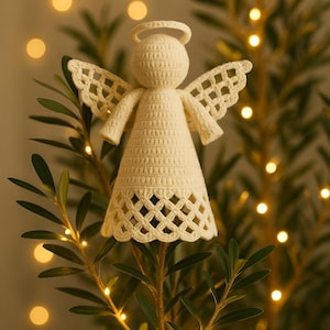 Puede incluir: Un ángel de ganchillo color crema, con un halo y alas caladas. El ángel está posado sobre una rama verde y frondosa, con luces suaves y desenfocadas en el fondo. Una decoración navideña festiva y hecha a mano.