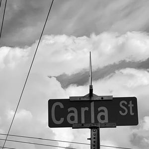 以下が含まれることがあります： モノクロ写真。雲の多い空を背景に、「Carla St.」と書かれた街路標識。電線と電柱がフレーム内に見え、都市の風景を補完しています。