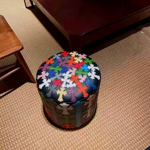 May include: A round, black ottoman with a colorful cross pattern on the top and sides. The crosses are in various colors, including red, orange, yellow, green, blue, and purple. The ottoman is a decorative furniture piece.